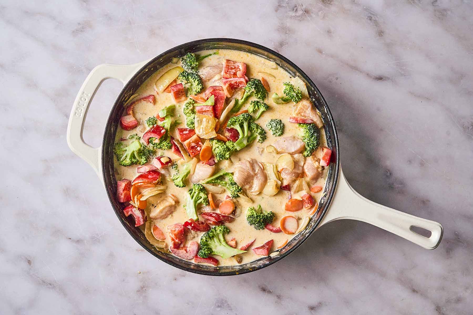 Chicken, red bell peppers, broccoli, and carrots simmering in a creamy yellow curry sauce in a cream-colored cast iron skillet.