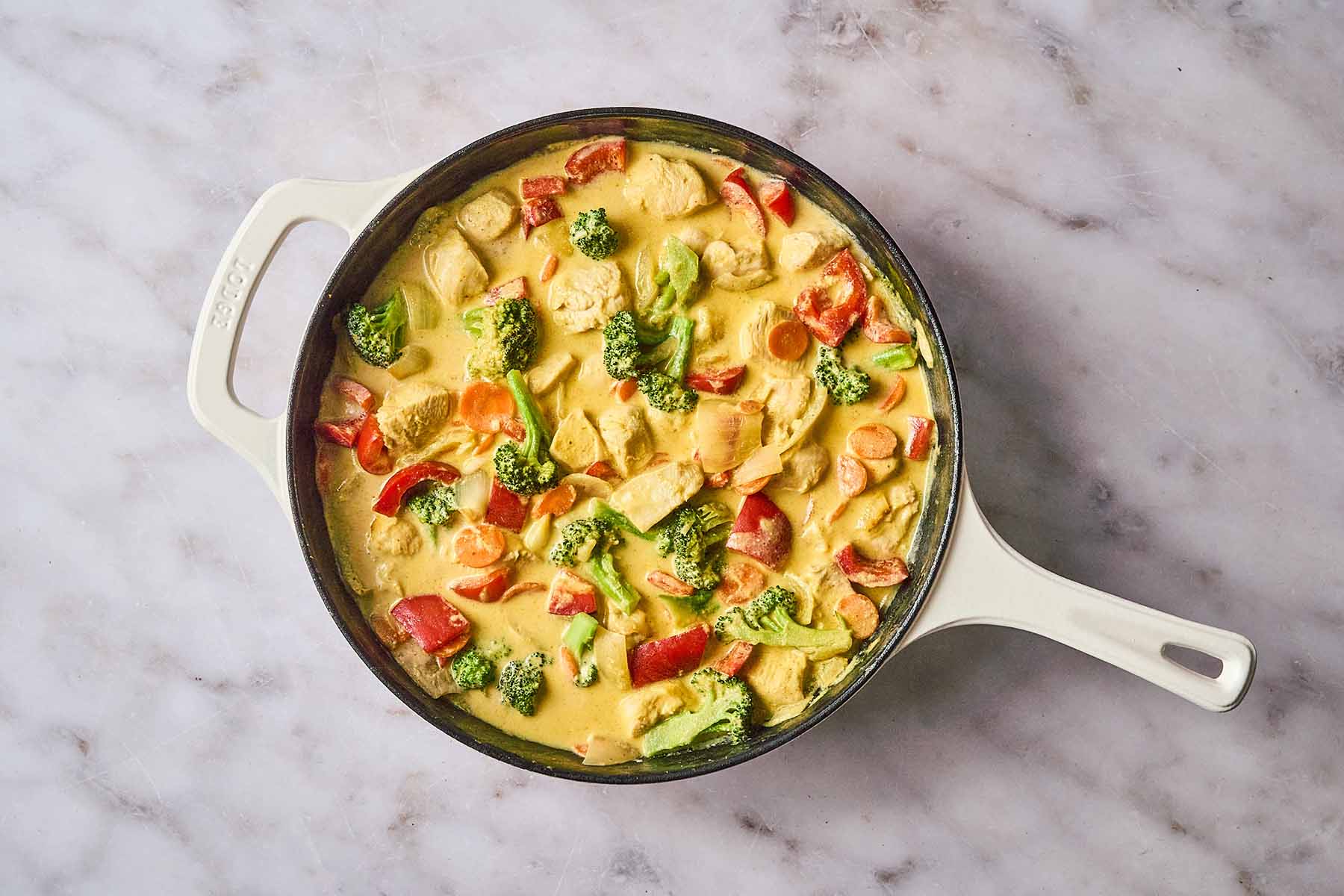 Fully cooked yellow curry chicken with red bell peppers, broccoli, and carrots simmering in a rich, creamy yellow curry sauce in a cream-colored cast iron skillet.