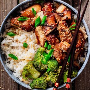 Top-down view of a bowl filled with teriyaki chicken thighs, white rice, and broccoli, garnished with sesame seeds and green onions, with chopsticks resting on the bowl.