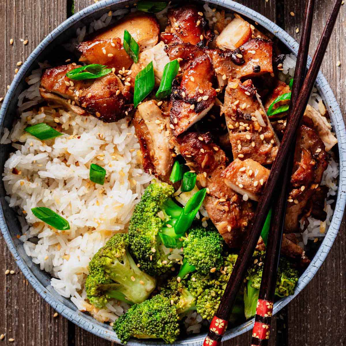 Top-down view of a bowl filled with teriyaki chicken thighs, white rice, and broccoli, garnished with sesame seeds and green onions, with chopsticks resting on the bowl.