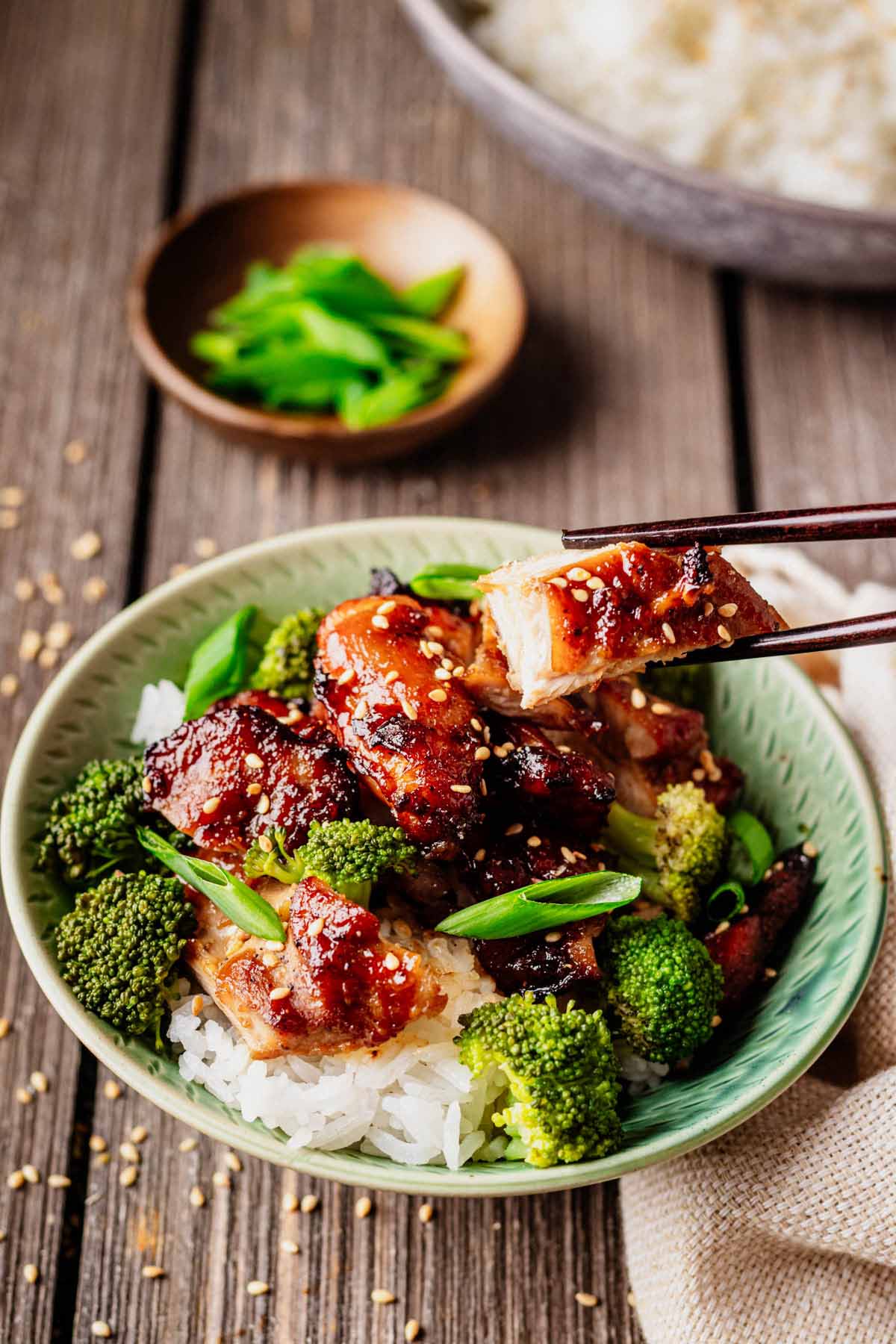 Green bowl filled with teriyaki chicken thighs, white rice, and broccoli, topped with sesame seeds and green onions, with chopsticks lifting a bite of glazed chicken.