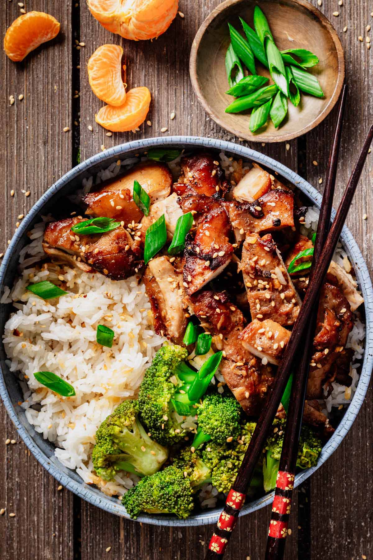 Bowl of air fryer teriyaki chicken thighs served over white rice with broccoli, garnished with sesame seeds and sliced green onions, with chopsticks resting on the side.
