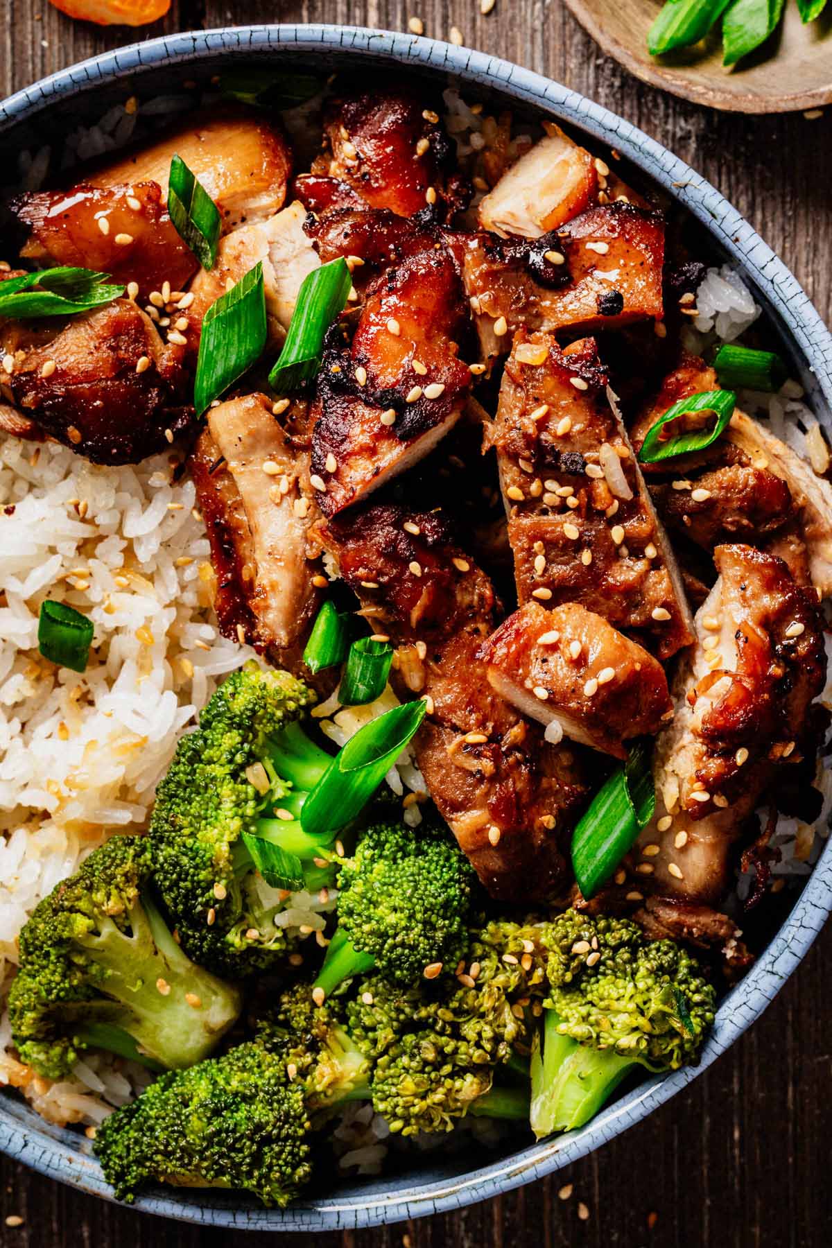 Close-up of sliced teriyaki chicken thighs served over white rice with steamed broccoli, garnished with sesame seeds and green onions in a blue ceramic bowl.
