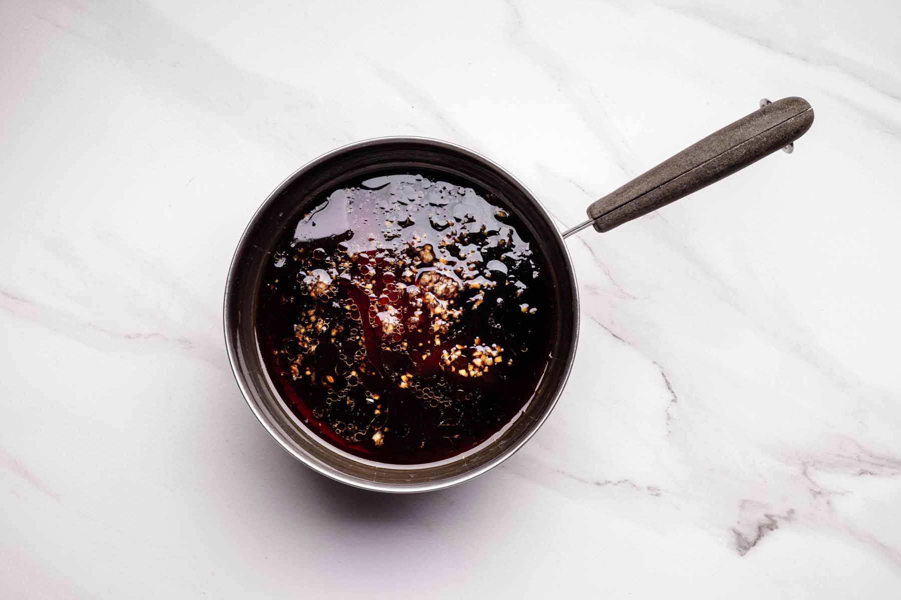 Small saucepan filled with dark teriyaki sauce, showing visible minced garlic and oil on the surface, sitting on a white marble countertop.