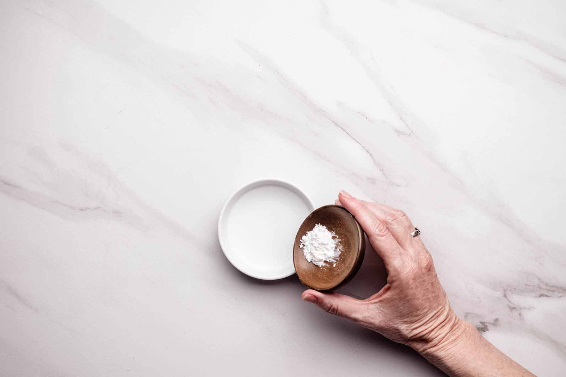 Hand holding a small wooden bowl of cornstarch above a white dish of water on a light marble countertop, preparing a slurry for thickening sauce.