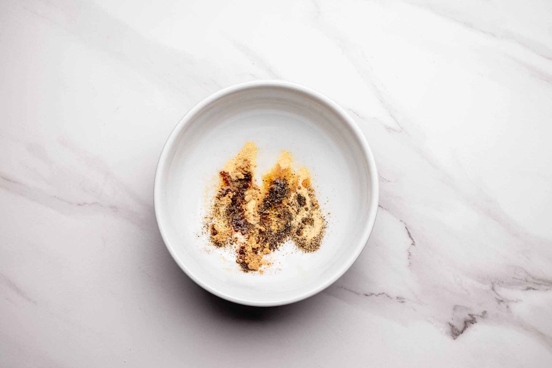 White bowl containing a mixture of dry spices and sesame oil, including garlic powder, ground ginger, and black pepper, on a light marble surface.