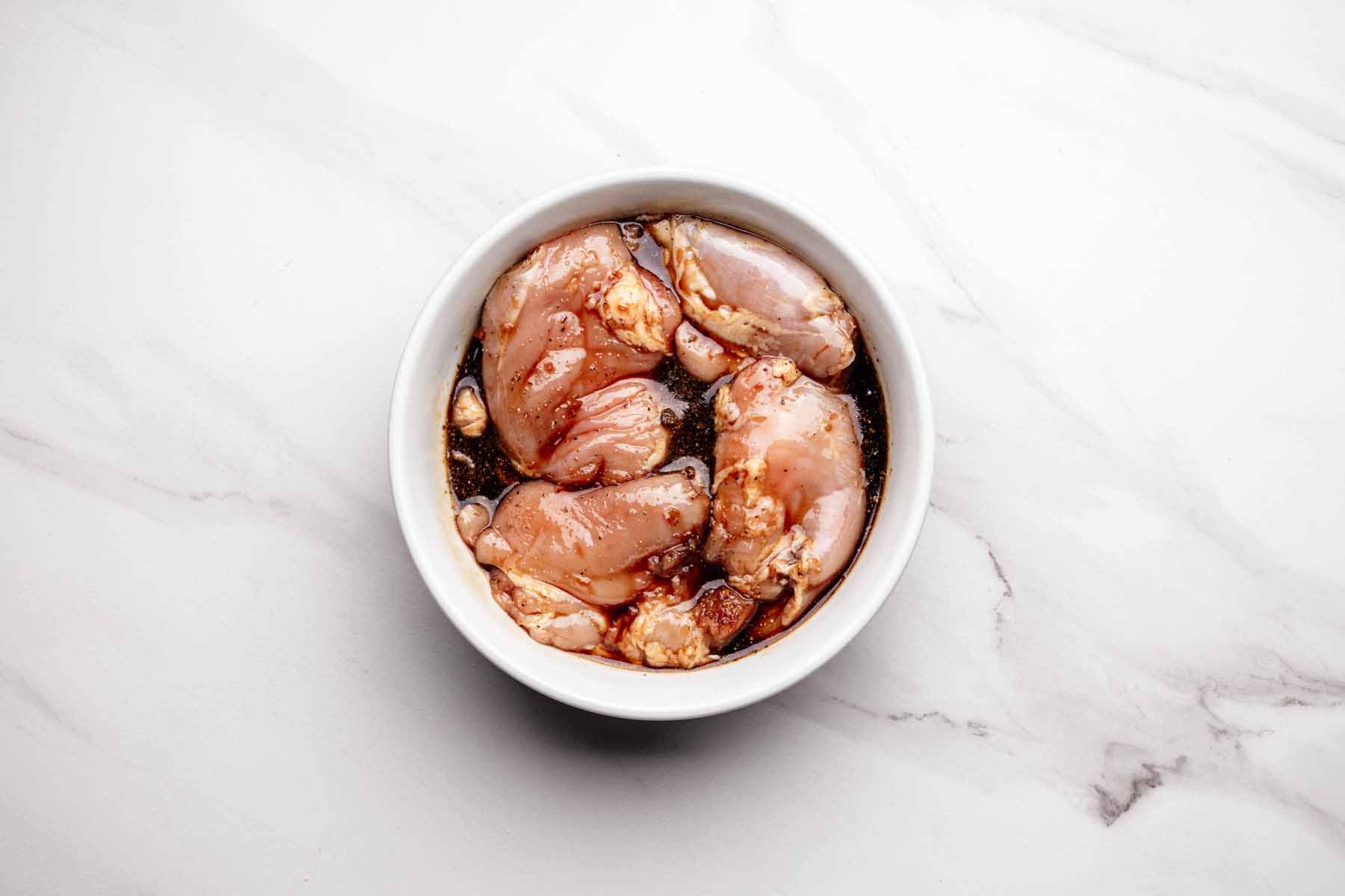 Raw boneless chicken thighs marinating in teriyaki sauce inside a white bowl, placed on a white marble surface.