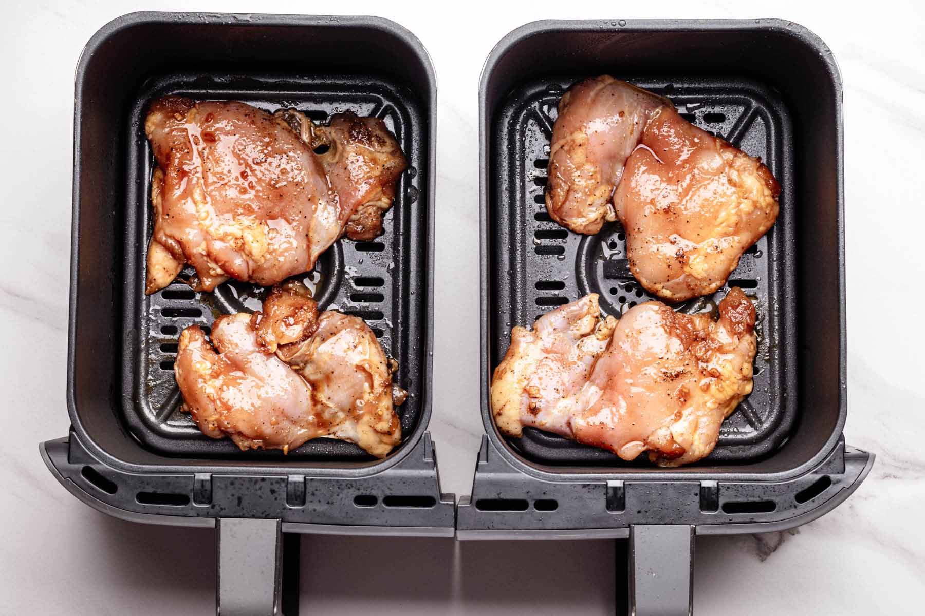 Raw marinated chicken thighs arranged in a double-basket air fryer, ready to be cooked, with visible seasoning and glossy teriyaki coating.