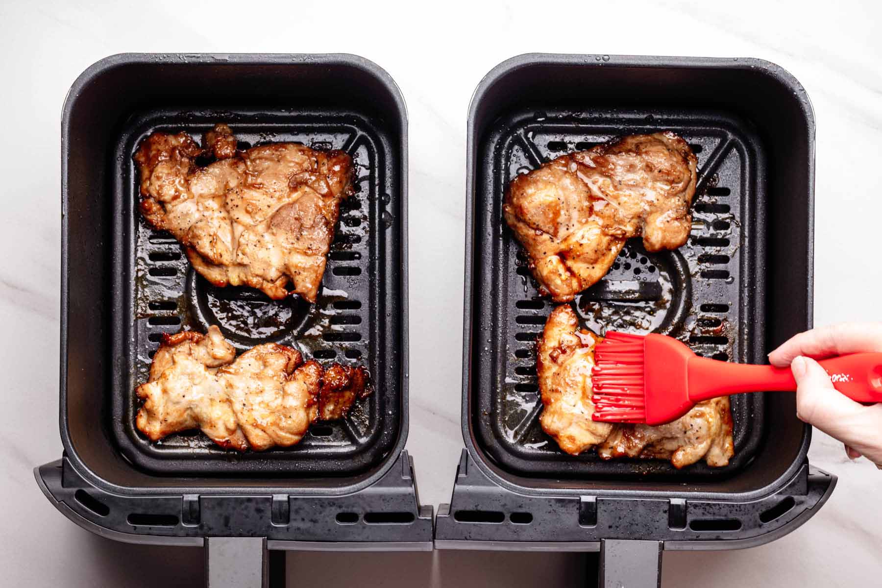 Partially cooked chicken thighs in a double-basket air fryer, with one piece being brushed with teriyaki sauce using a red silicone basting brush.