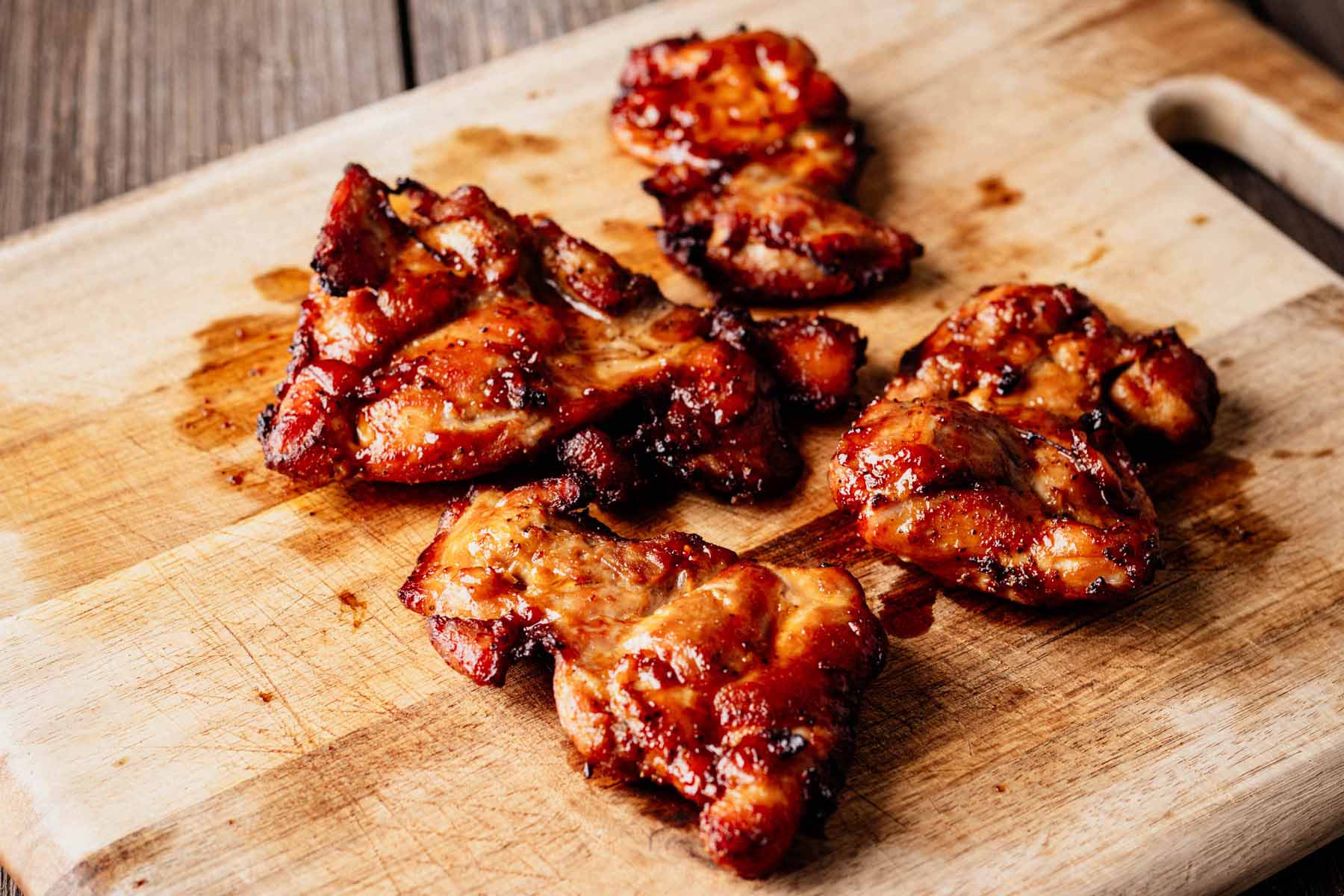 Cooked teriyaki chicken thighs resting on a wooden cutting board, showing a glossy, caramelized glaze and slightly charred edges.