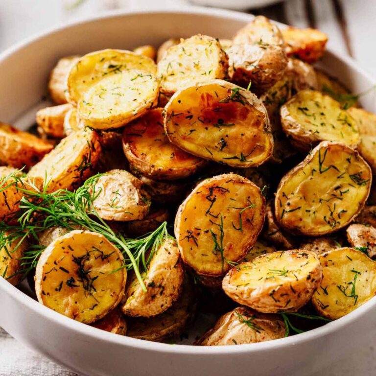 Close-up of golden, crispy dill roasted potatoes in a white bowl, garnished with fresh dill.