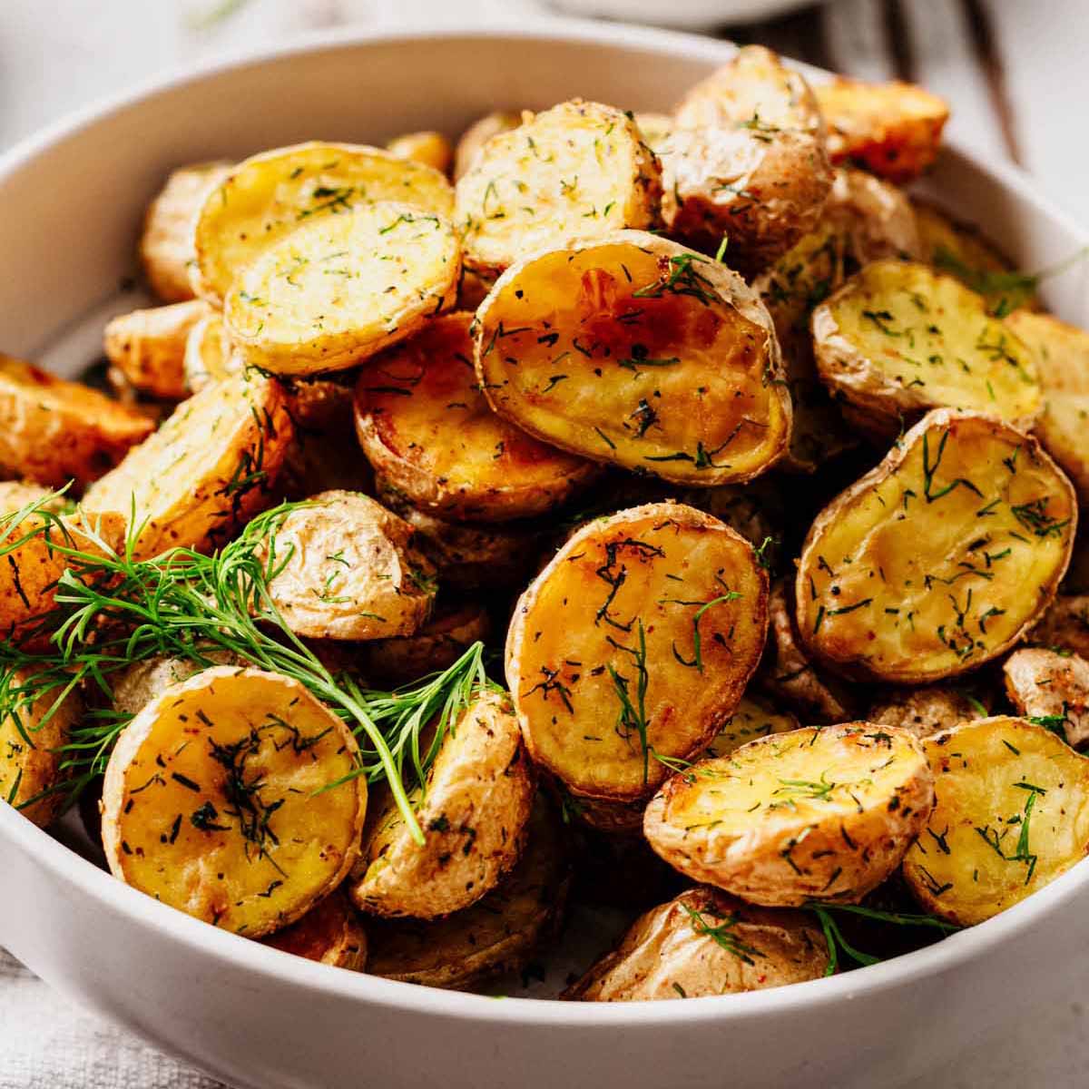 Close-up of golden, crispy dill roasted potatoes in a white bowl, garnished with fresh dill.