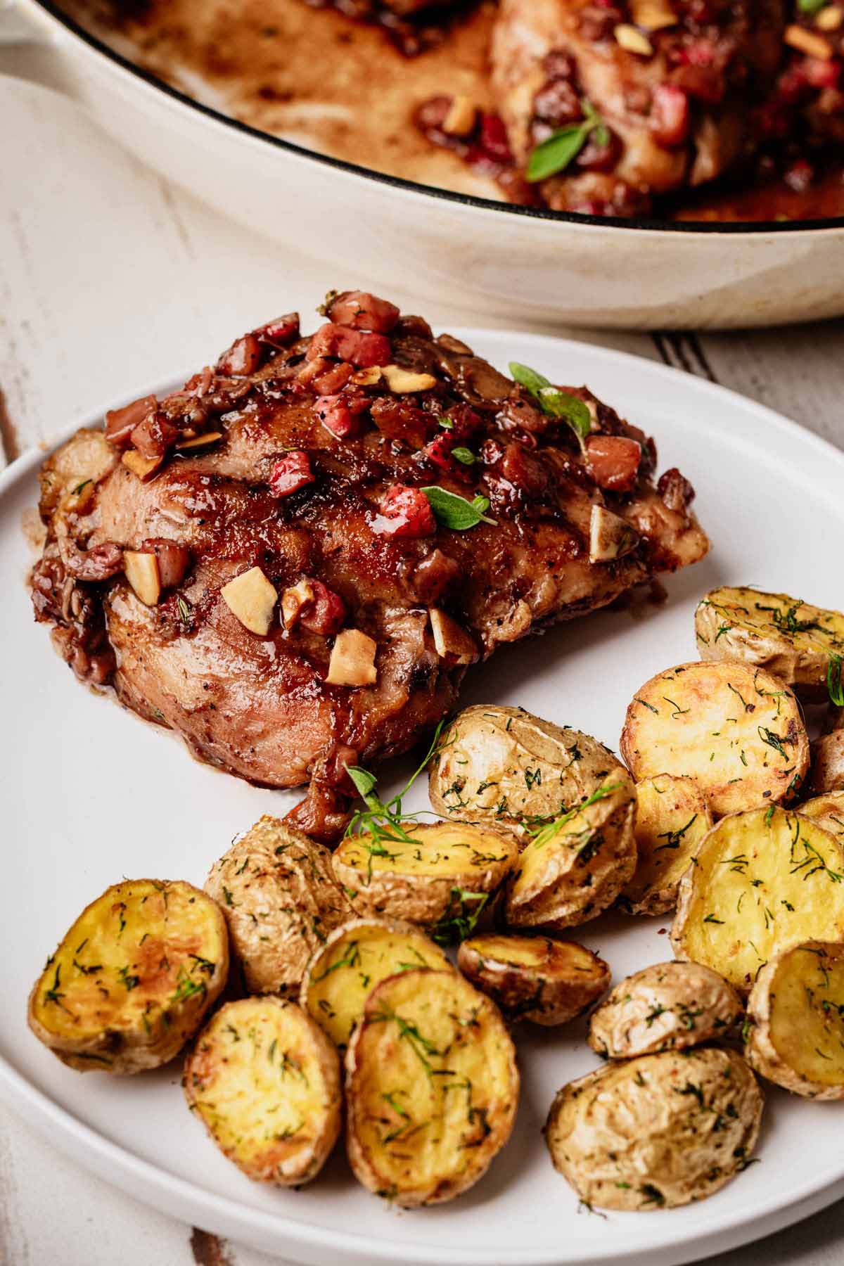 Dill roasted potatoes served alongside a glazed pork chop topped with apple and bacon, plated on a white dish.