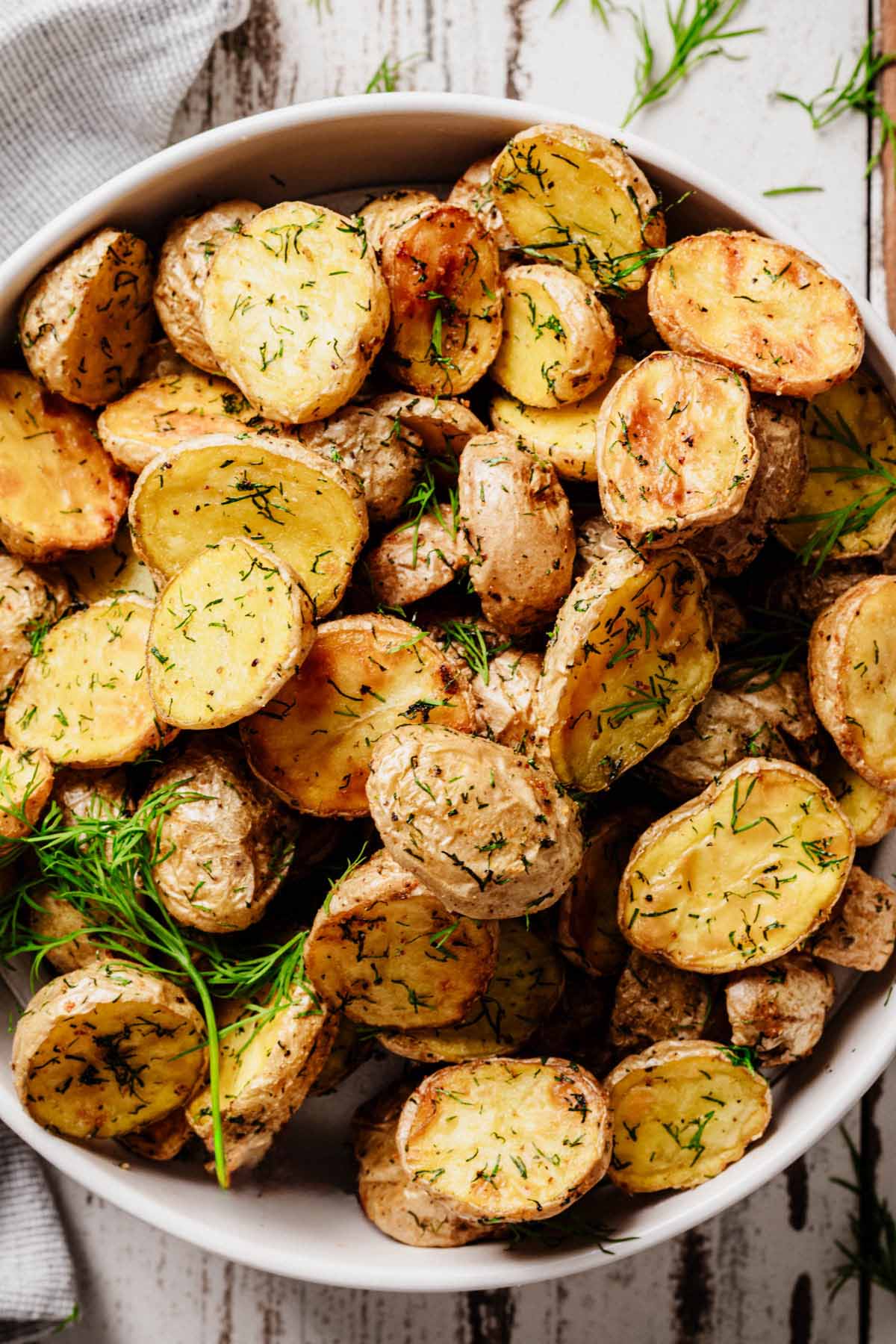 Golden roasted baby potatoes with crispy edges and fresh dill, served in a white bowl on a rustic surface.
