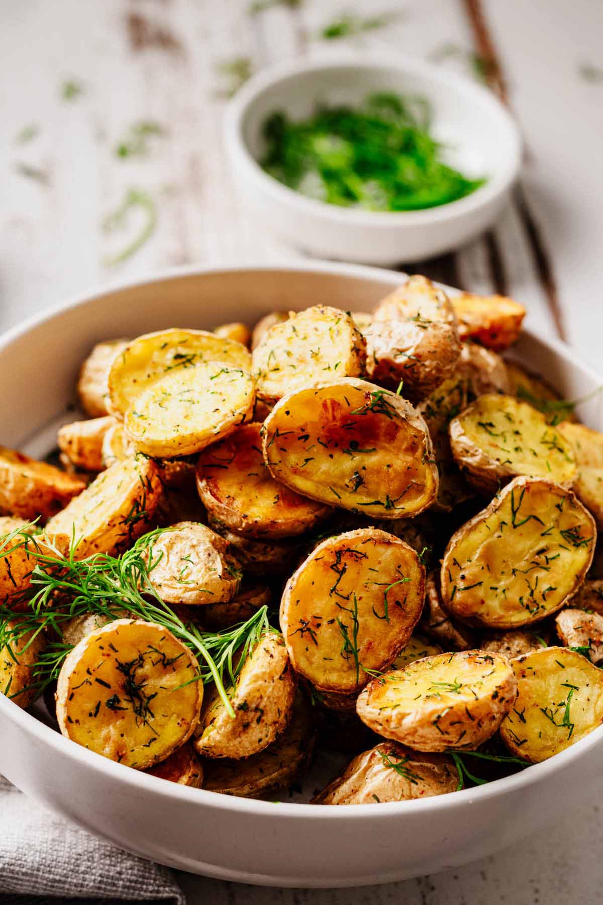 Close-up of golden roasted baby potatoes with fresh dill, served in a white bowl with a small dish of herbs in the background.