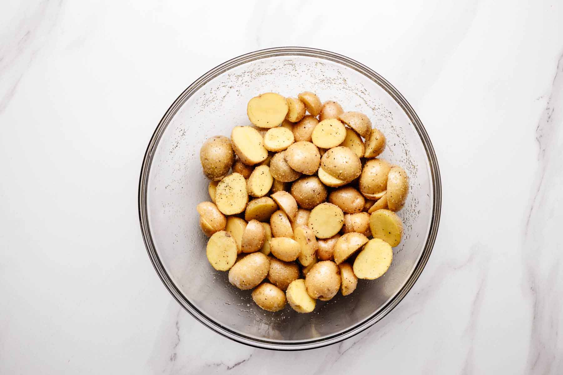 Halved baby potatoes tossed with oil, salt, pepper, and garlic powder in a glass bowl.