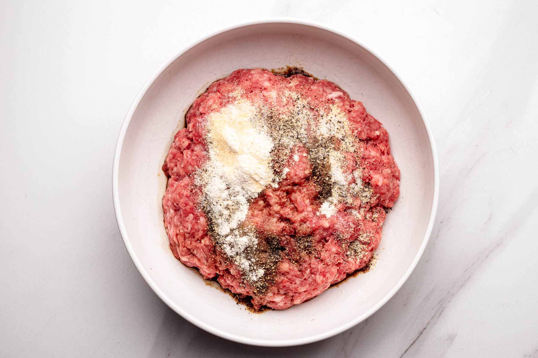 Ground beef in a white bowl topped with Worcestershire sauce, salt, pepper, garlic powder, and onion powder, ready to be mixed for slider patties.
