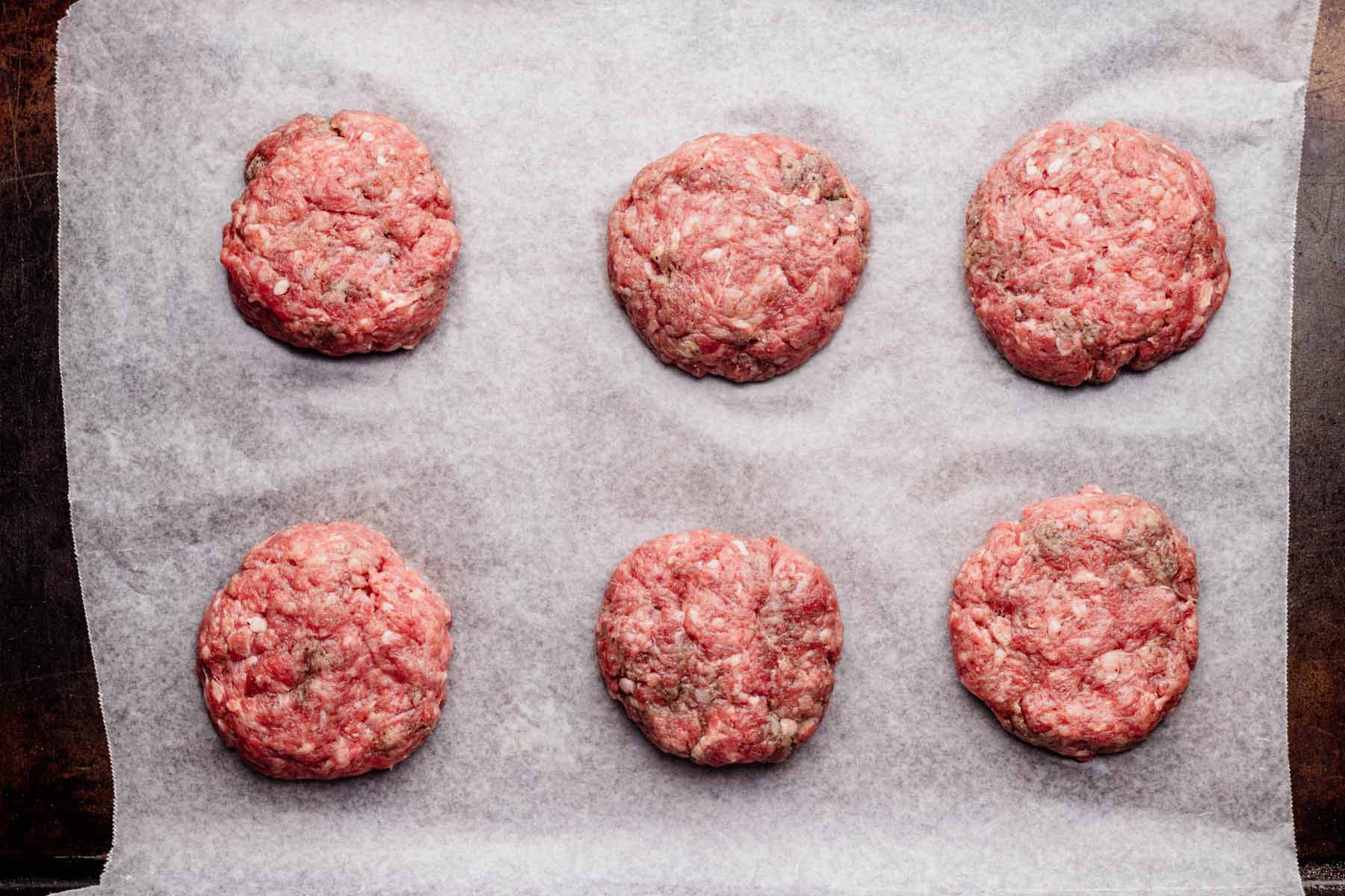 Six raw slider-sized beef patties arranged on a parchment-lined baking sheet, ready for cooking.