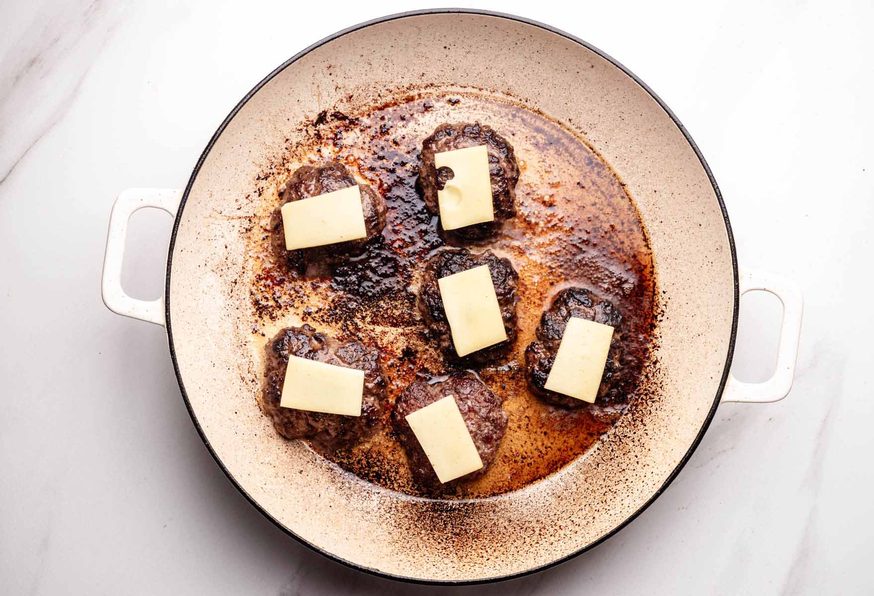 Six cooked beef slider patties in a skillet, each topped with a slice of Gruyère cheese before melting.
