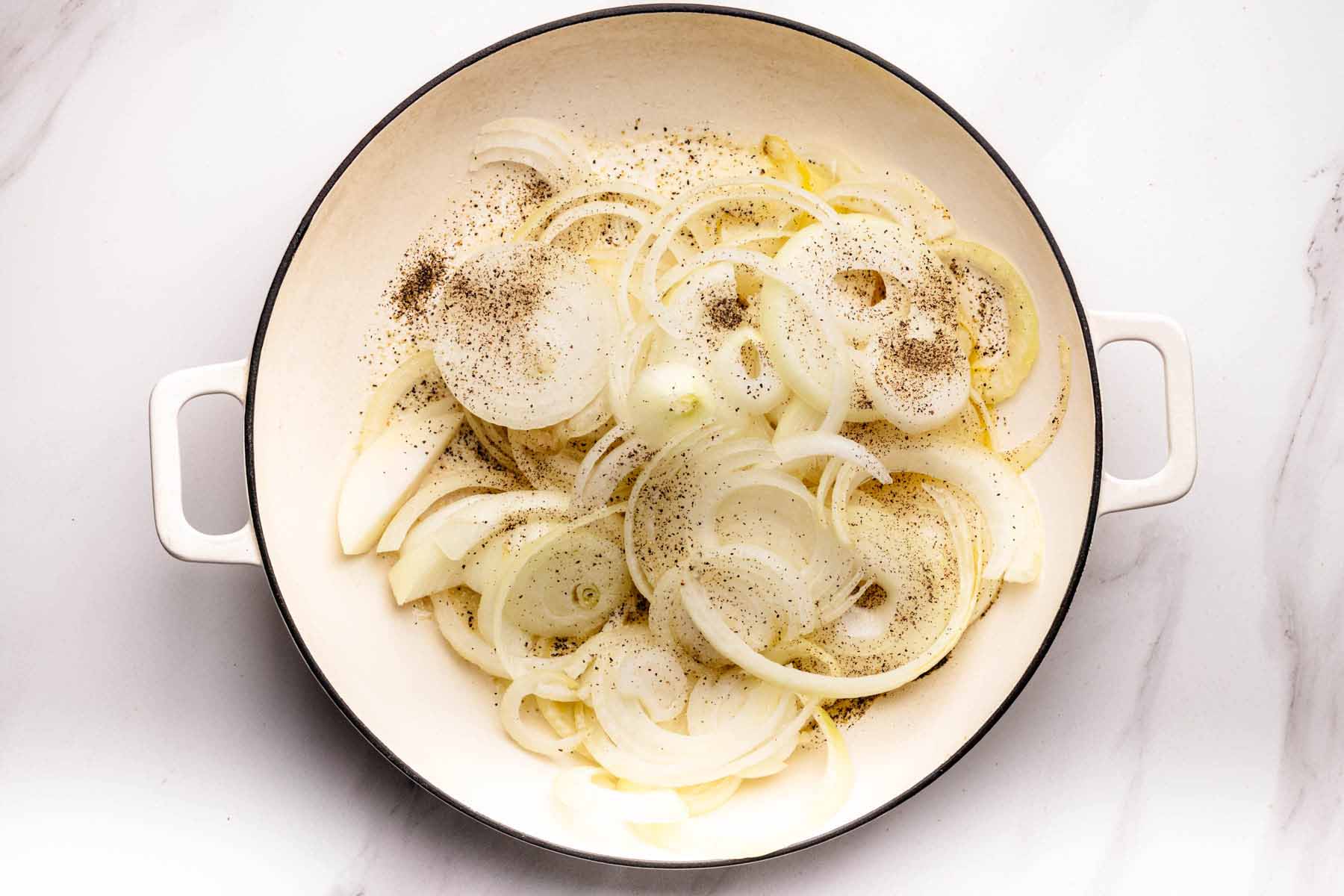 Thinly sliced onions with salt and pepper in a white skillet, ready to be caramelized.