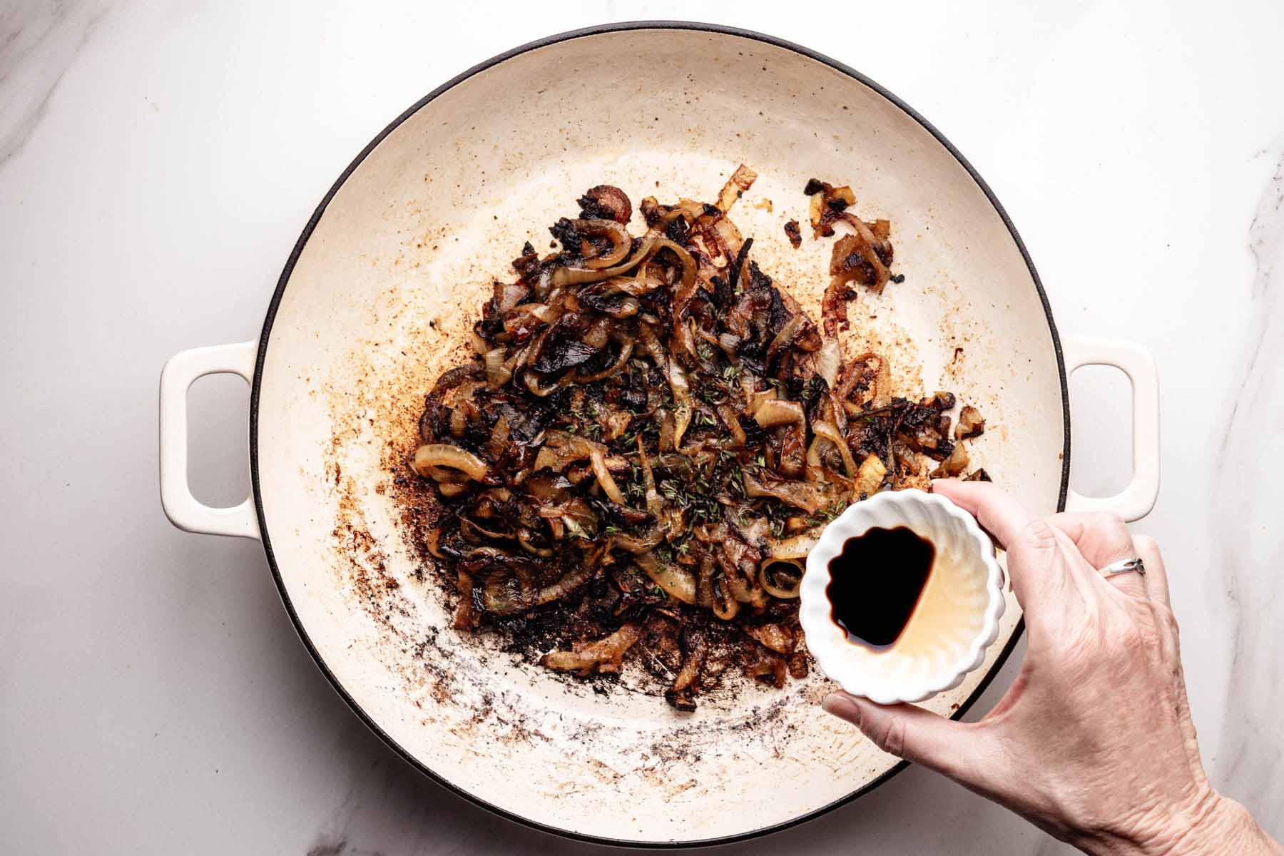 Caramelized onions with thyme in a skillet, with a hand holding a small dish of balsamic vinegar about to pour it in.