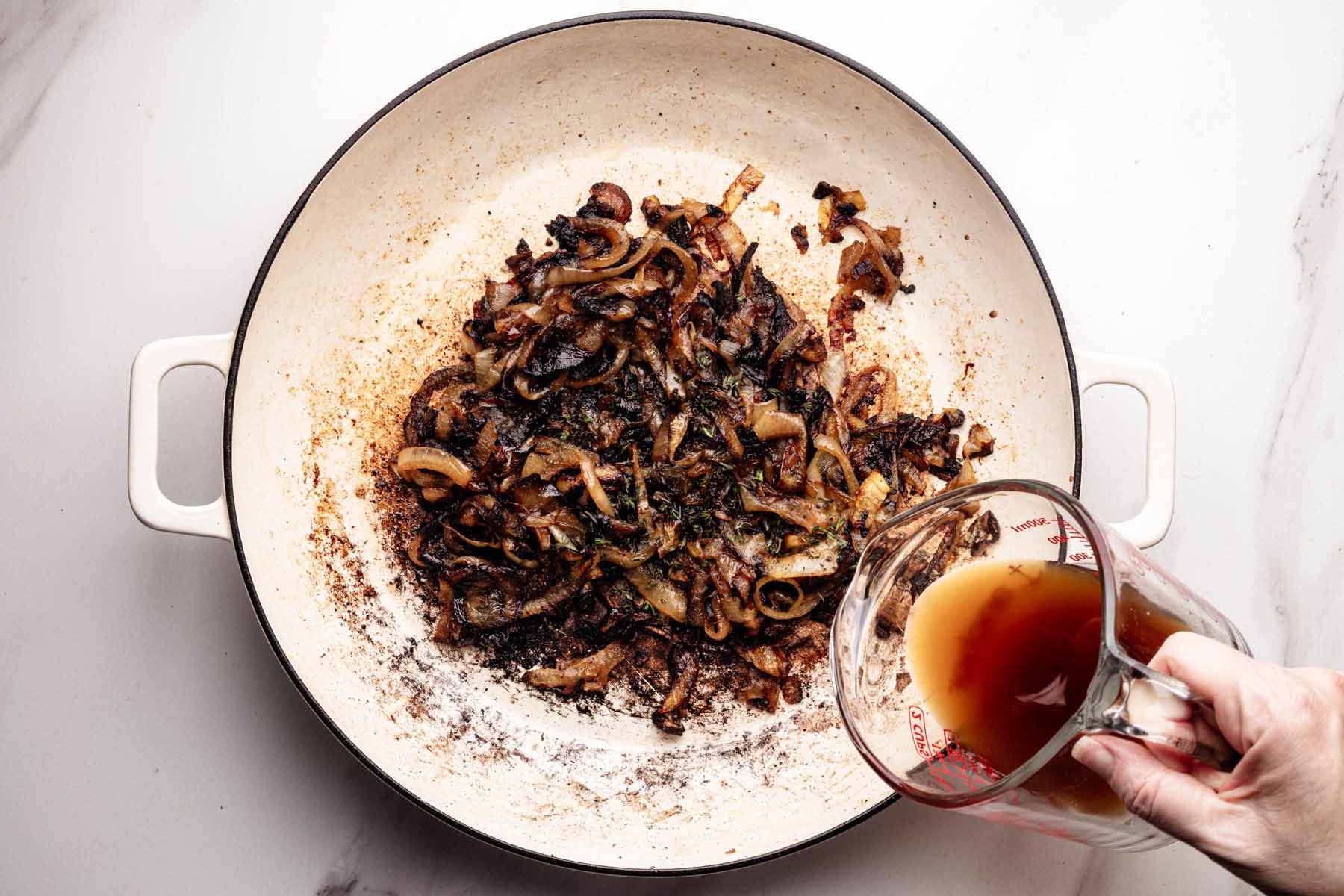 Caramelized onions in a skillet with a hand pouring beef broth from a measuring cup to deglaze the pan.