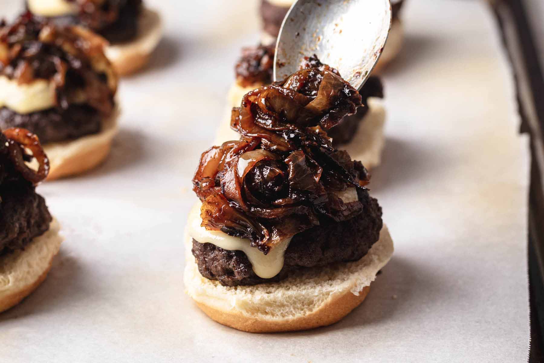 A spoonful of caramelized onions being added to a slider with melted cheese and a beef patty on the bottom bun.