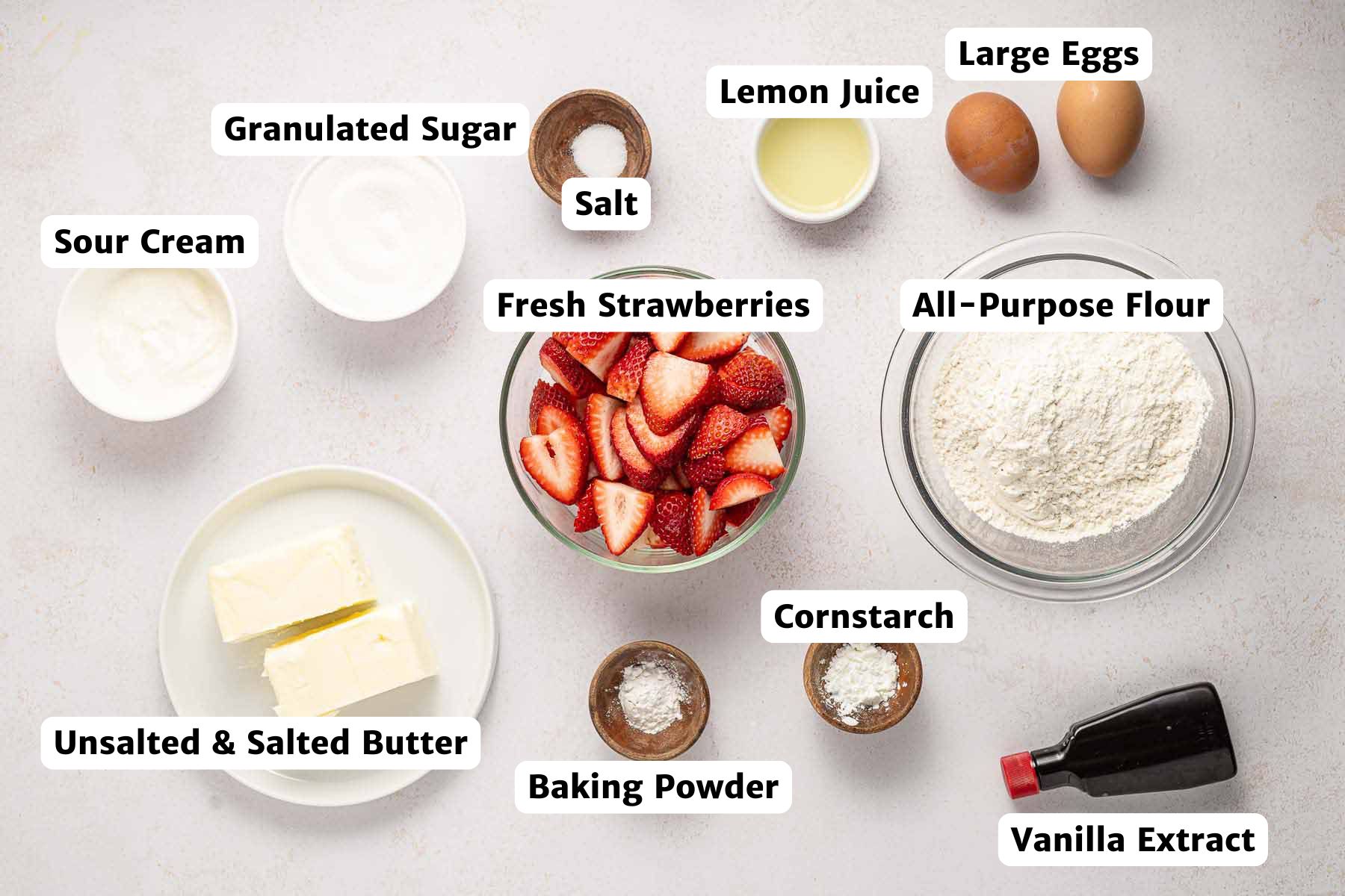 Labeled flat lay of ingredients for strawberry upside down cake, including fresh strawberries, flour, sugar, eggs, butter, sour cream, lemon juice, vanilla extract, baking powder, cornstarch, and salt