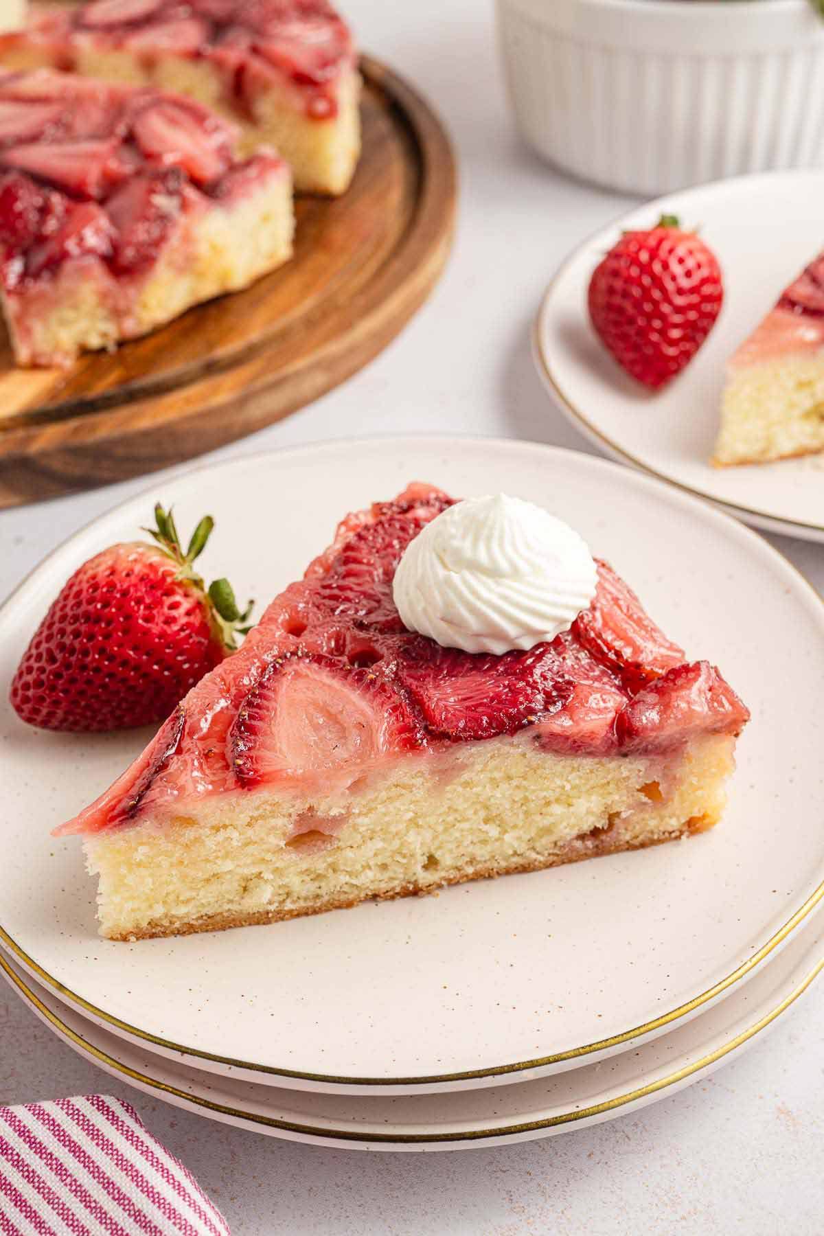 Slice of strawberry upside down cake topped with whipped cream, served on a white plate with a fresh strawberry on the side