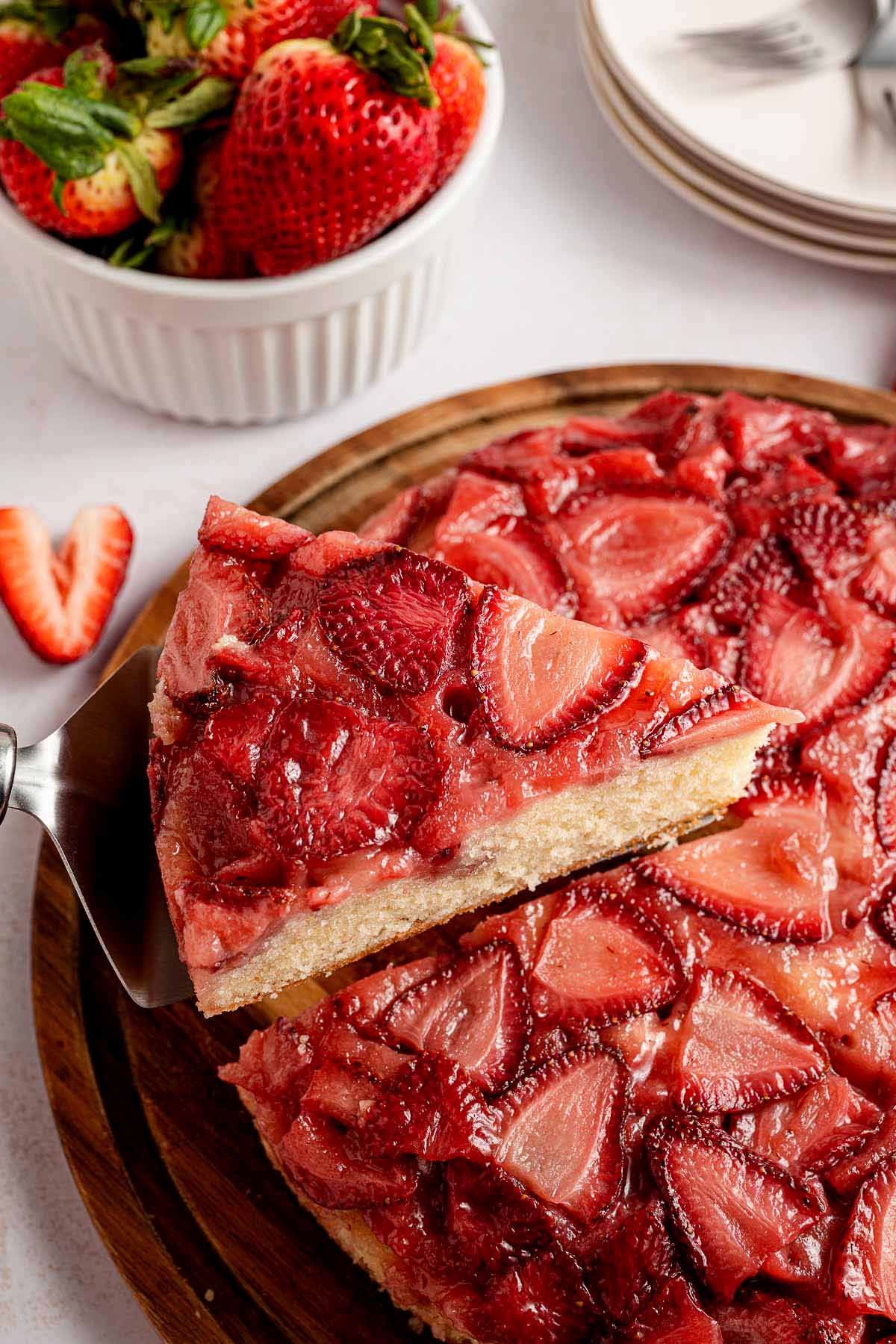 Close-up of a slice of strawberry upside down cake being lifted from a wooden serving board, with fresh strawberries and stacked dessert plates in the background.