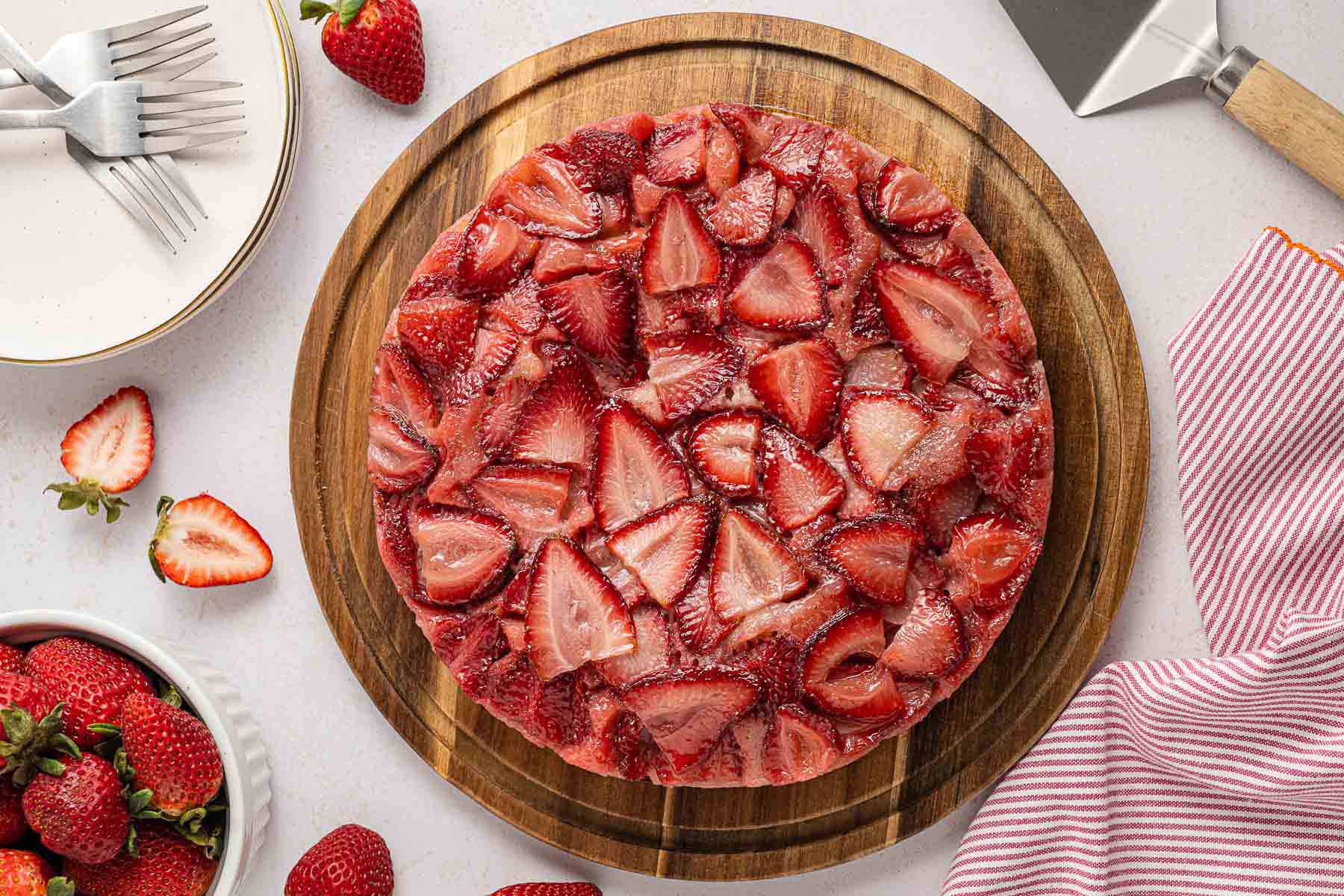 Inverted strawberry upside down cake on a wooden board, topped with glossy baked strawberries and ready to serve with plates, forks, and fresh berries nearby
