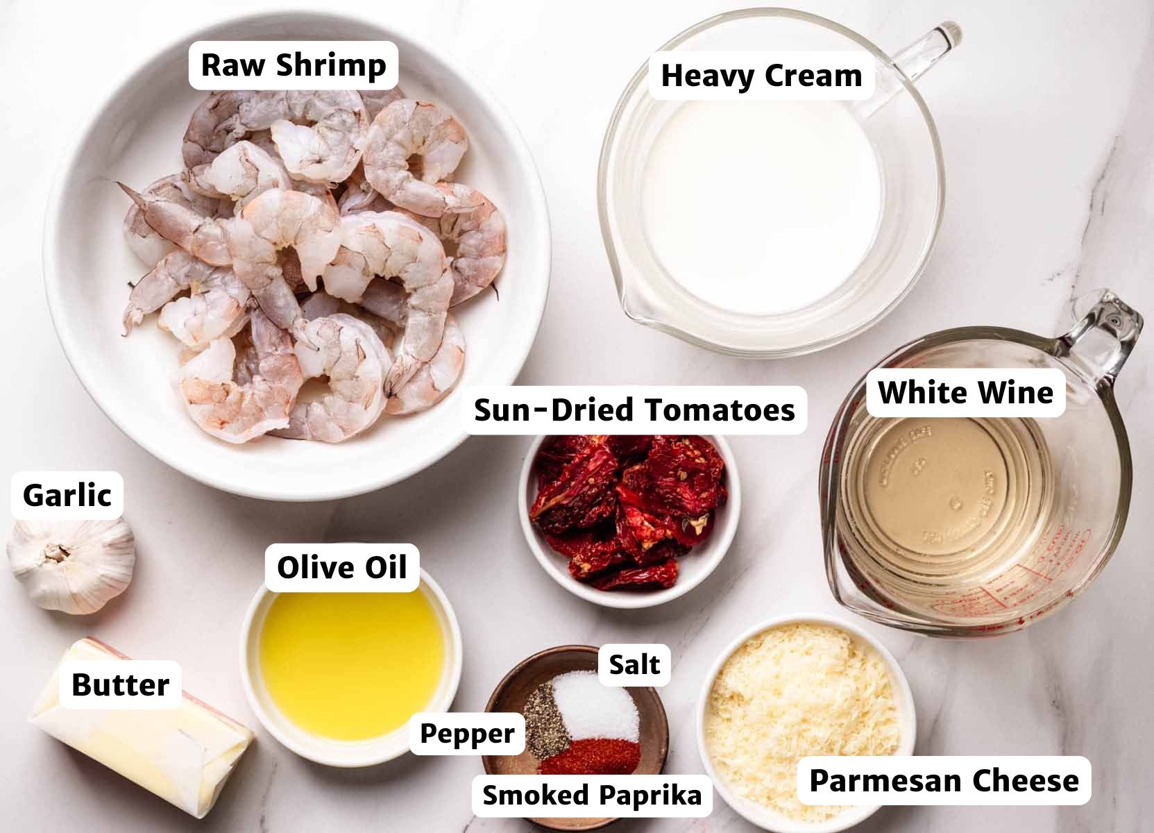 Overhead view of labeled ingredients for sun-dried tomato shrimp, including raw shrimp in a white bowl, heavy cream, white wine, sun-dried tomatoes, Parmesan cheese, olive oil, butter, garlic, and small bowls of salt, pepper, and smoked paprika.