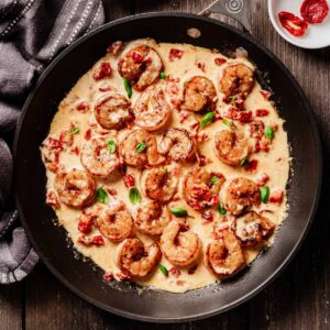 Overhead view of sun-dried tomato shrimp in a creamy sauce, garnished with fresh basil and served in a black skillet on a rustic wooden table.