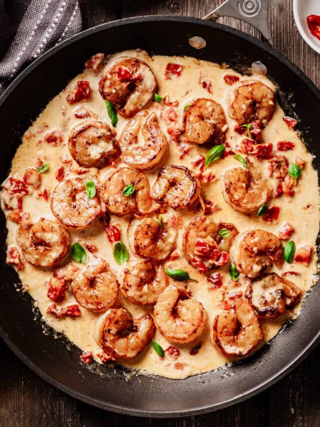 Overhead view of sun-dried tomato shrimp in a creamy sauce, garnished with fresh basil and served in a black skillet on a rustic wooden table.