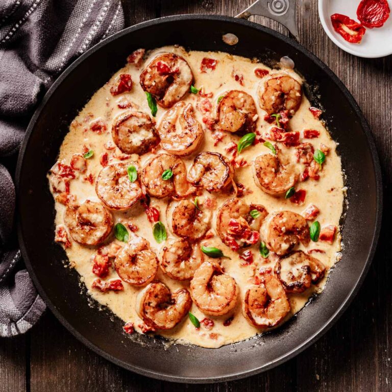 Overhead view of sun-dried tomato shrimp in a creamy sauce, garnished with fresh basil and served in a black skillet on a rustic wooden table.