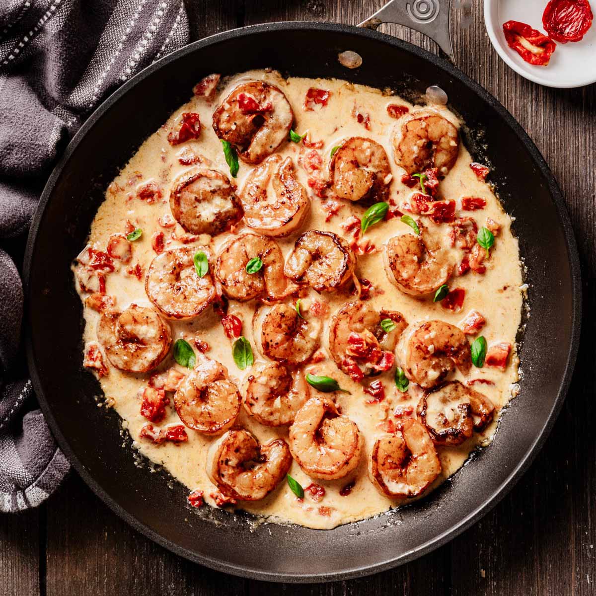 Overhead view of sun-dried tomato shrimp in a creamy sauce, garnished with fresh basil and served in a black skillet on a rustic wooden table.