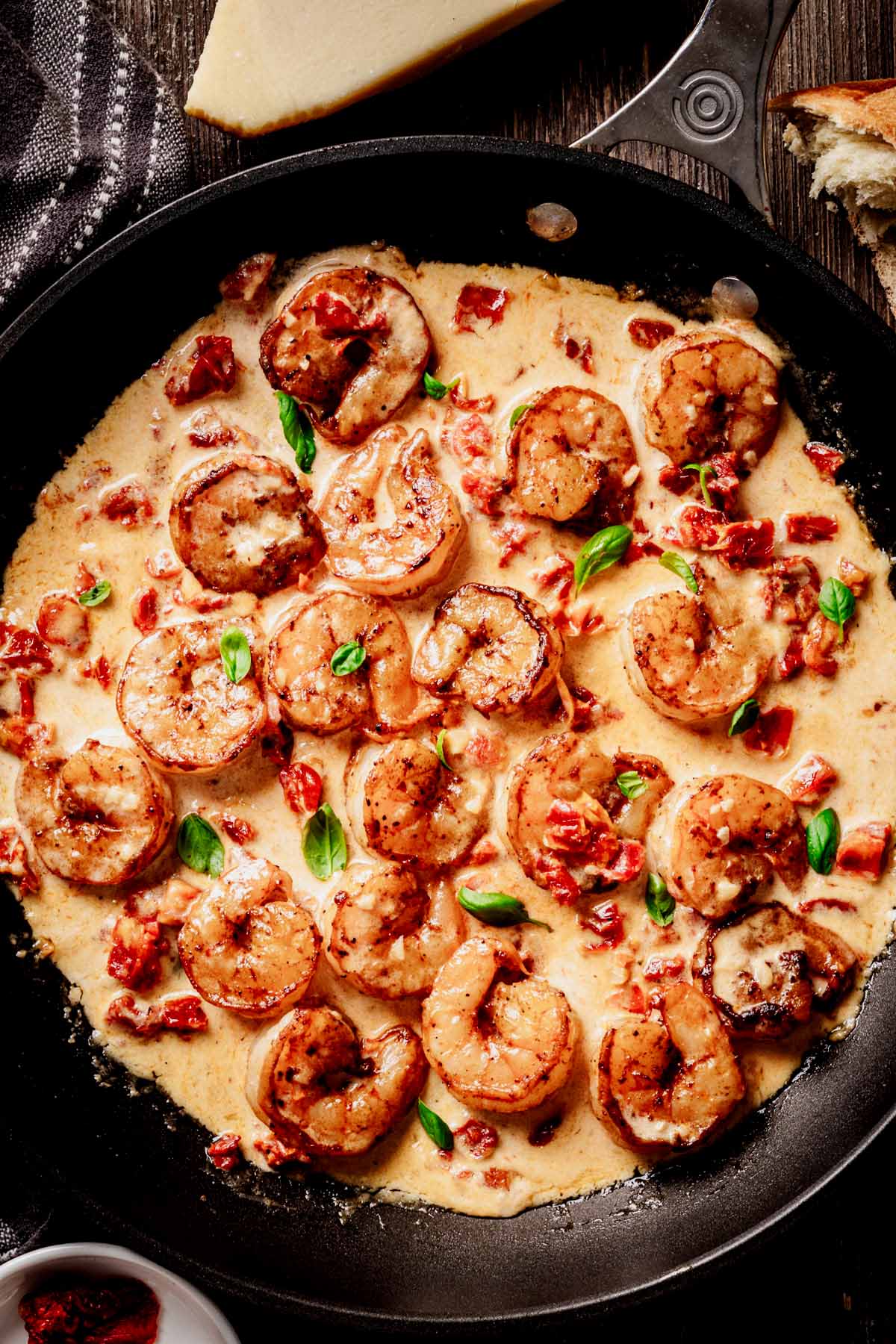 Close-up overhead view of creamy sun-dried tomato shrimp in a skillet, garnished with fresh basil, with crusty bread and Parmesan wedge visible on the side.