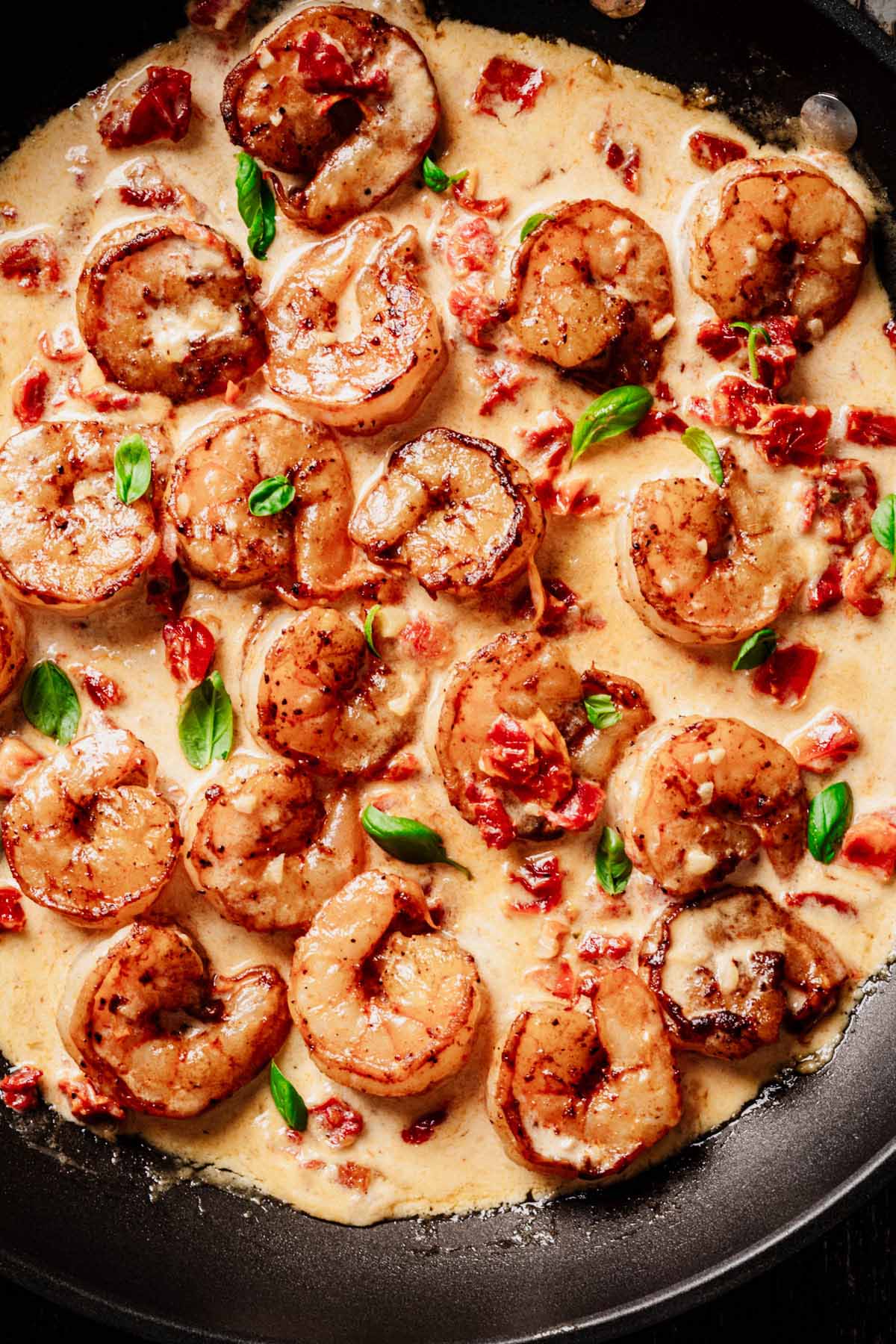 Close-up overhead view of shrimp coated in a creamy sun-dried tomato sauce, garnished with fresh basil, in a black skillet.