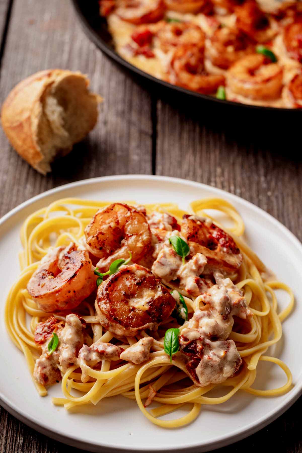 Close-up of creamy sun-dried tomato shrimp served over linguine on a white plate, garnished with fresh basil, with crusty bread and skillet in the background on a wooden table.