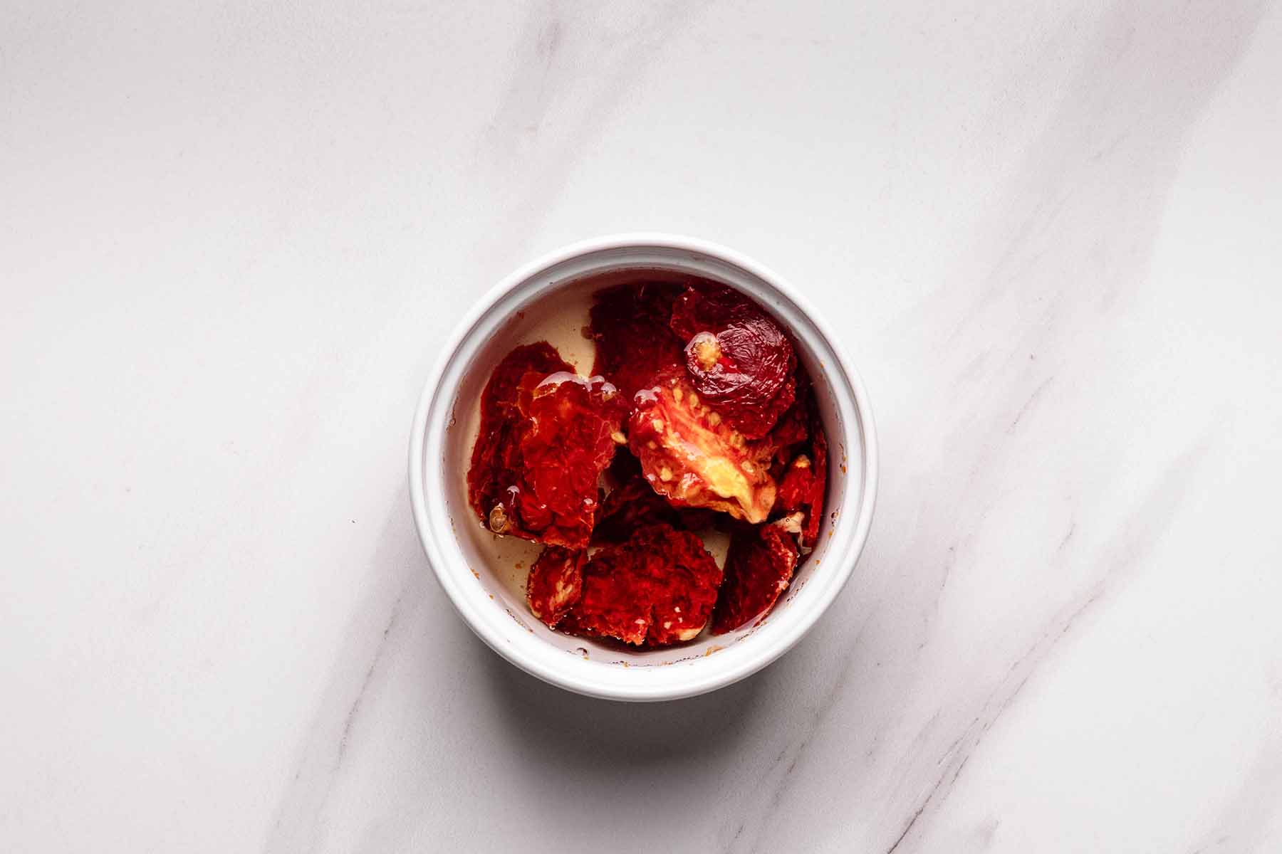 Overhead view of a white bowl filled with sun-dried tomatoes soaking in hot water to rehydrate, placed on a light marble surface.