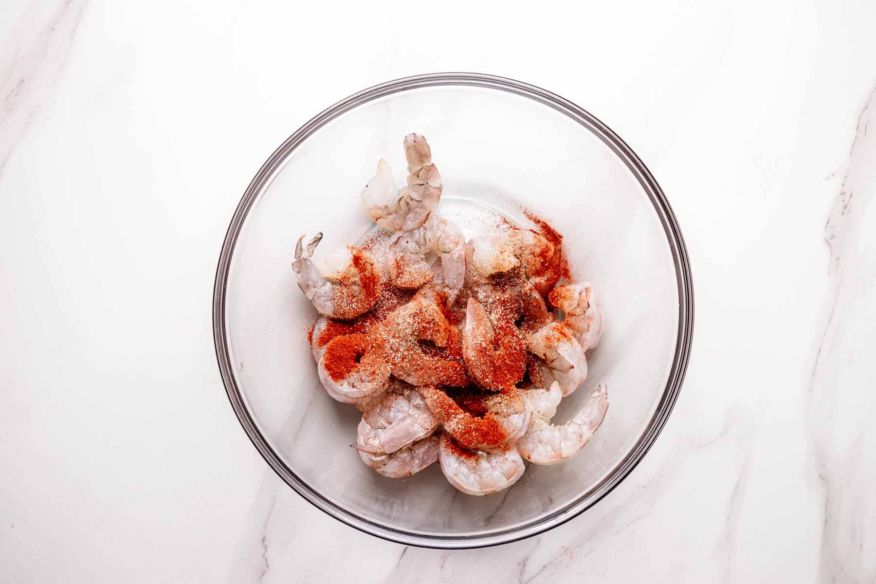 Overhead view of raw shrimp in a glass bowl seasoned with salt, pepper, and smoked paprika, ready to be tossed before cooking.