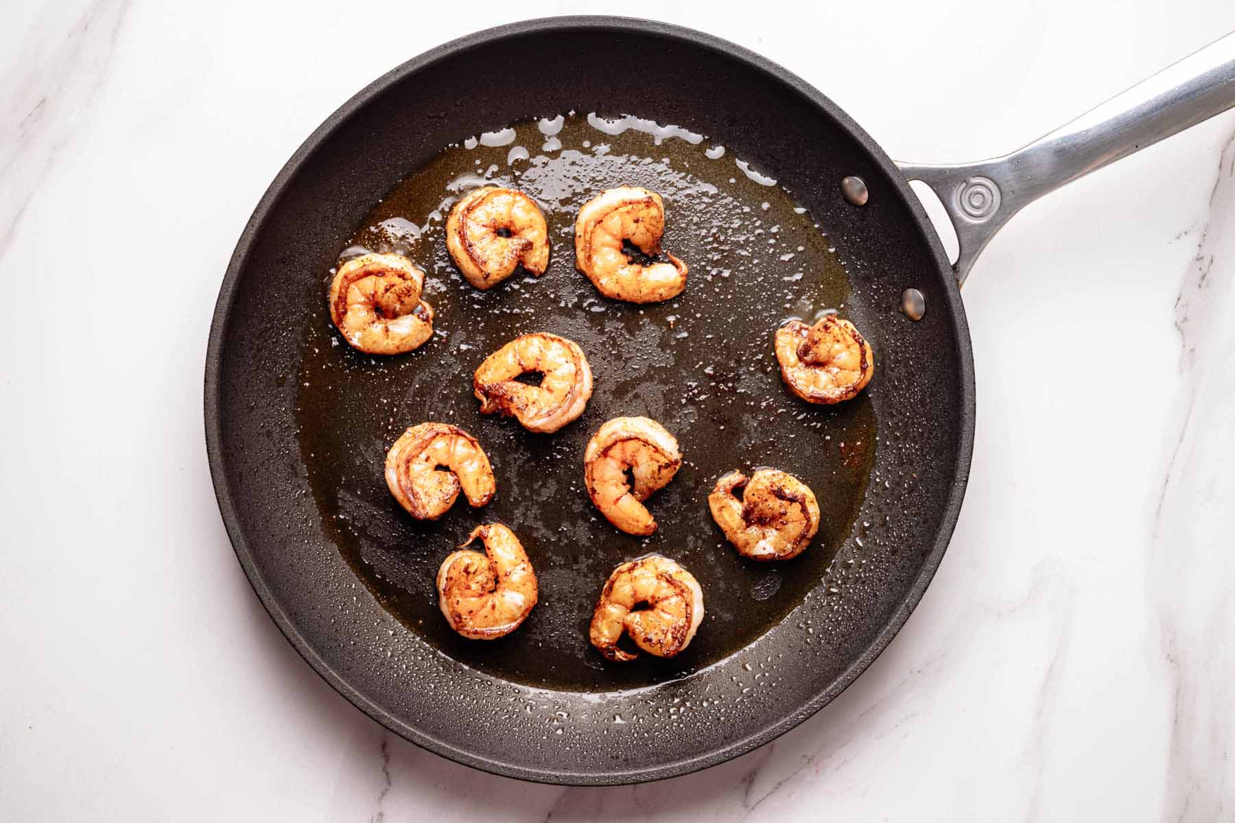 Overhead view of seared shrimp cooking in a skillet with olive oil, showing golden edges and slight charring.