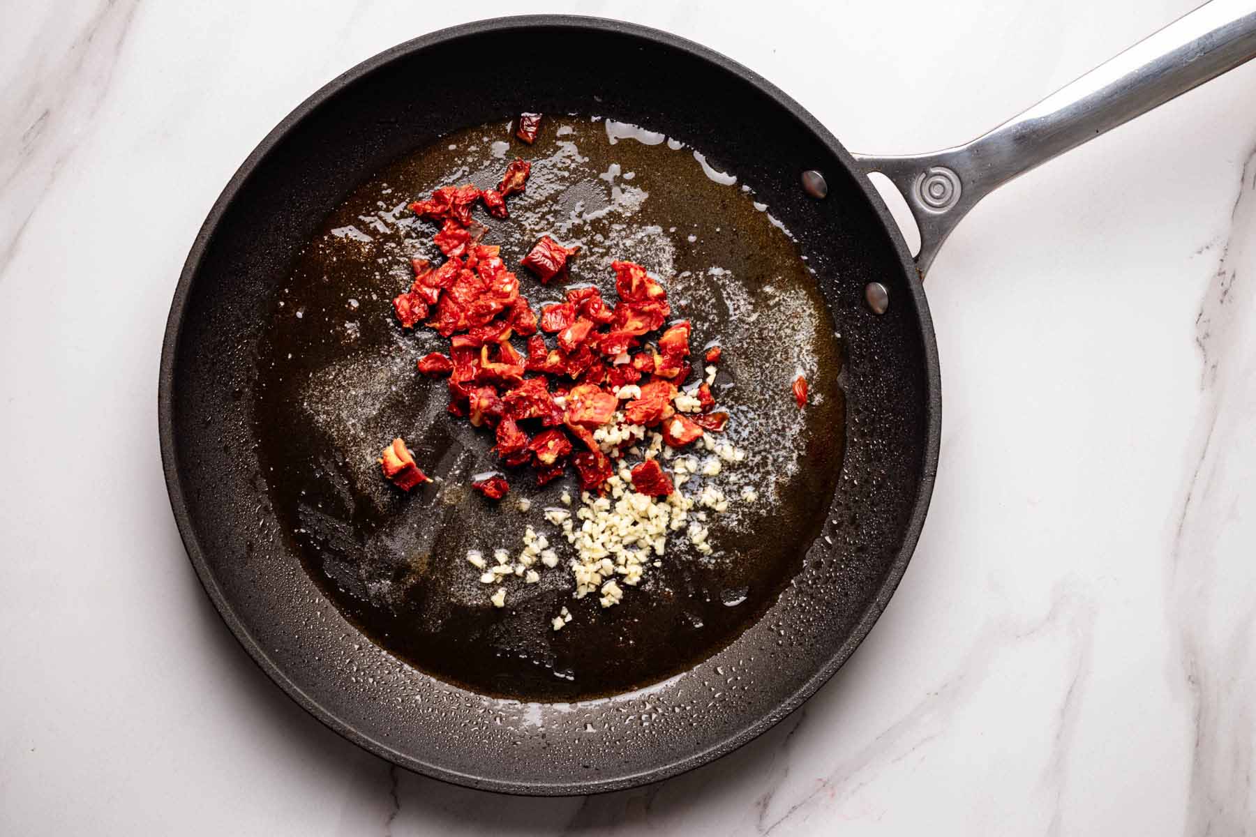 Overhead view of a skillet with chopped sun-dried tomatoes and minced garlic sautéing in melted butter and shrimp drippings.