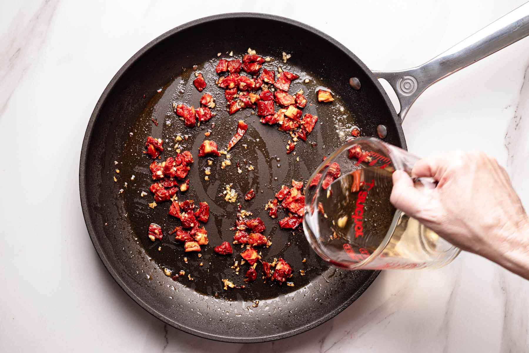 Overhead view of chopped sun-dried tomatoes and garlic in a skillet as white wine is being poured in from a glass measuring cup to deglaze the pan.