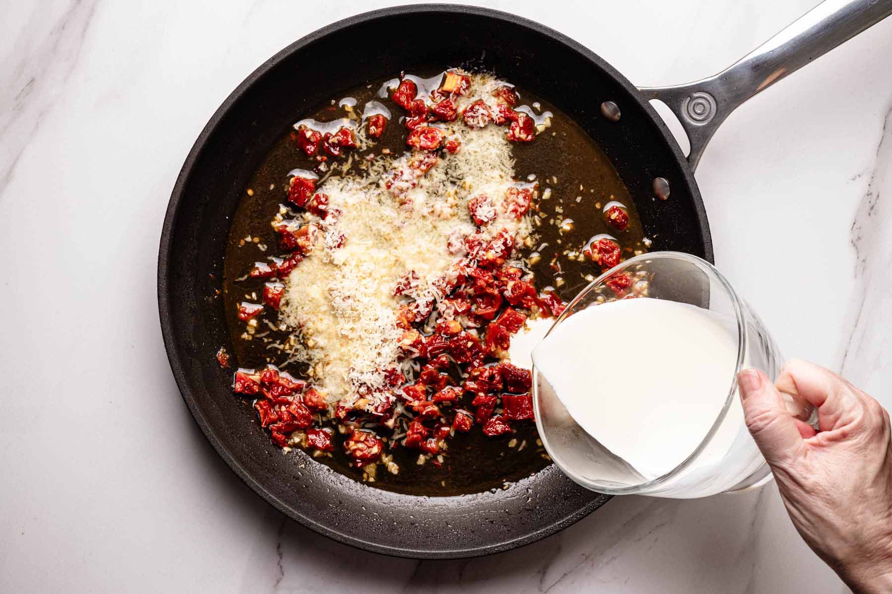 Overhead view of a skillet with sun-dried tomatoes, garlic, and grated Parmesan as heavy cream is being poured in to create a creamy sauce.