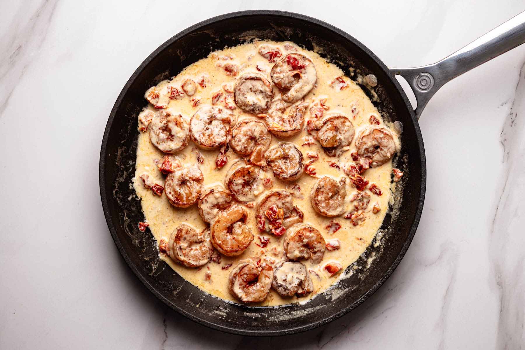 Overhead view of shrimp simmering in a creamy sun-dried tomato sauce in a skillet, with the sauce thickened and clinging to the shrimp.