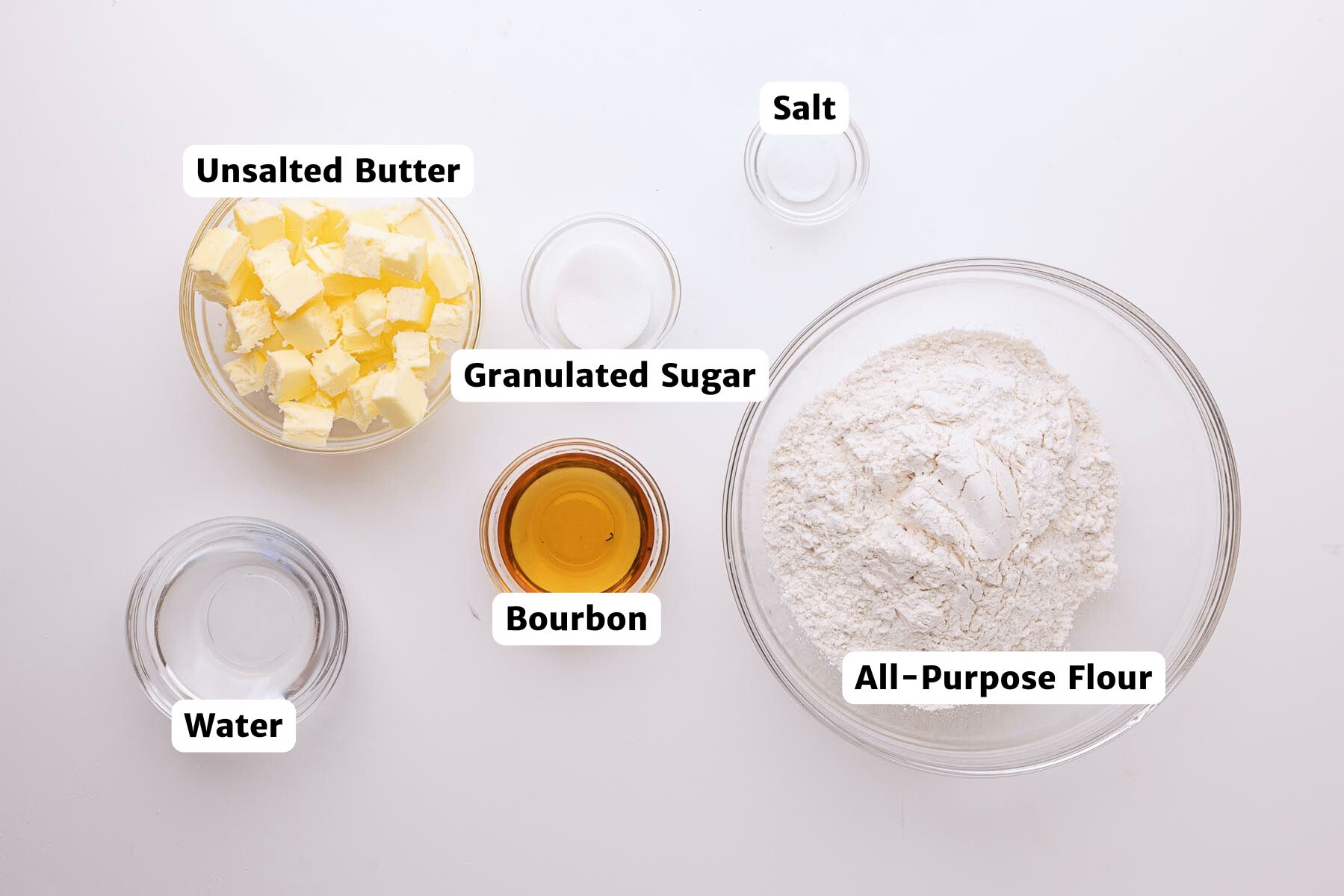 Labeled ingredients for bourbon butter double pie crust, including cubed unsalted butter, granulated sugar, salt, all-purpose flour, bourbon, and ice water arranged in glass bowls on a white surface.