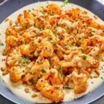 Close-up of harissa roasted cauliflower with whipped cottage cheese, honey-tahini drizzle, toasted pine nuts, and fresh herbs, served on a blue plate.
