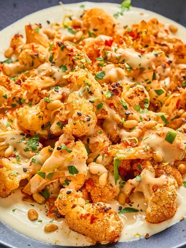 Close-up of harissa roasted cauliflower with whipped cottage cheese, honey-tahini drizzle, toasted pine nuts, and fresh herbs, served on a blue plate.