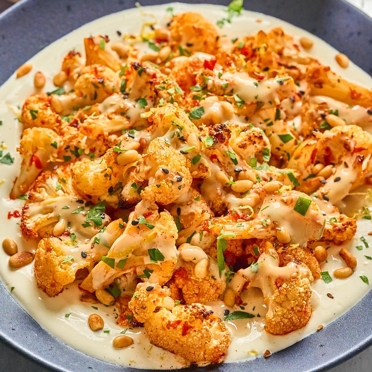 Close-up of harissa roasted cauliflower with whipped cottage cheese, honey-tahini drizzle, toasted pine nuts, and fresh herbs, served on a blue plate.